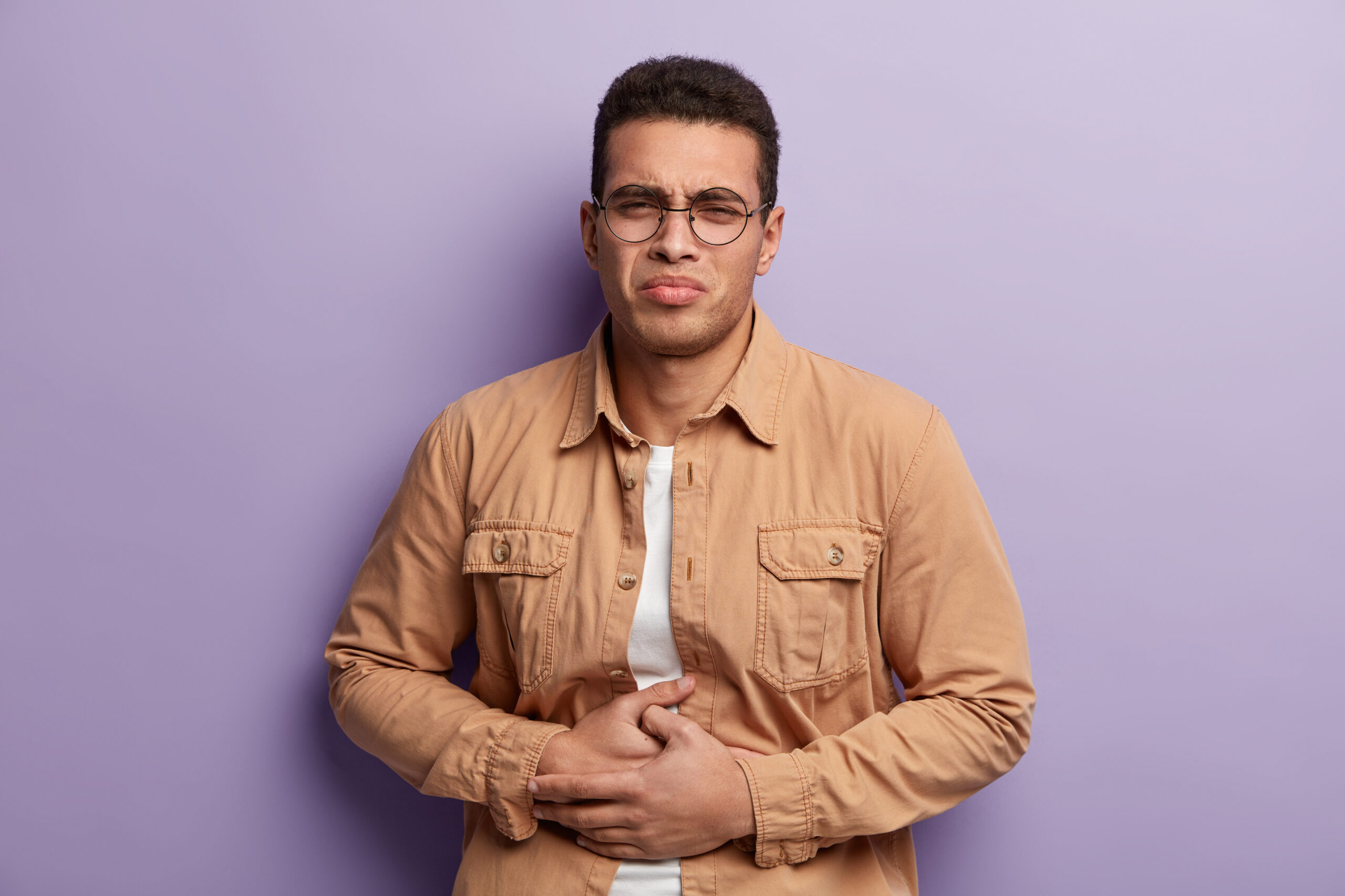 Upset male with dark short hair, suffers from stomach pain, keeps hands on belly, regrets eating fast food, has painful feelings and discomfort, wears beige shirt, isolated over purple background.